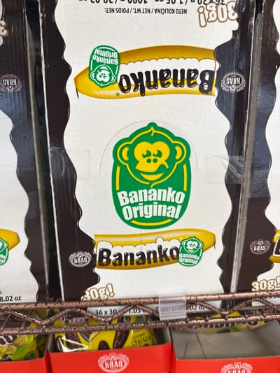 The image shows stacked boxes of "Bananko Original" chocolate-covered banana candies on a metal wire shelf, likely in a retail environment. The packaging is white with a yellow banana graphic, the brand name "Bananko" in black letters, and a green logo with a monkey. The boxes have brown tops and scalloped edges. The brand "KRAS" is also visible on the boxes. Text on the boxes indicates the product weight "30g," and "NETO KOLIČINA/NET WT./POIDS NET: 1080g/38.02 oz." The shelf is likely in a grocery store, possibly in Darien, USA. A red box is seen to the right of the Bananko boxes.