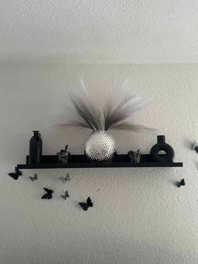 A black shelf is mounted on a textured white wall. On the shelf are a black bottle, a small decorative container, a silver vase with wispy grey and white decorative plumes, another small decorative container, and a black circular vase with a hole in the center. Below the shelf, black and patterned butterfly decals are affixed to the wall. The scene appears to be an interior decoration. This image was captured in Bielefeld, Germany.