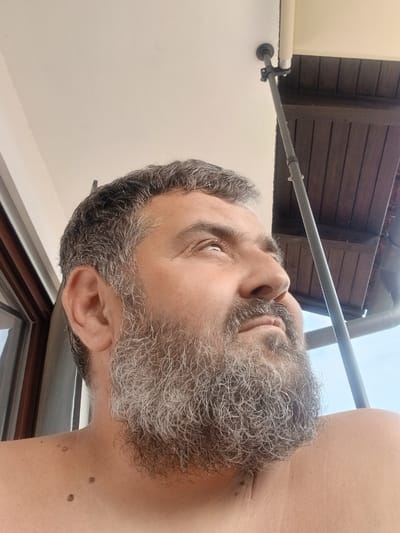 The media file is a close-up, downward-angled selfie of a man with graying hair and beard, taken outdoors. The man's face, shoulder, and part of his chest are visible. He has several moles on his shoulder and upper chest. His right eye is open and looking towards the camera, while his left eye is closed or mostly obscured. The lighting suggests it is daytime, possibly morning or afternoon, with bright natural light.
In the background, a dark brown wooden ceiling or overhang is visible, and a portion of a white roller blind or screen. A gray pole extends across the image from the left foreground towards the right background, attached to the ceiling by a bracket. The overall scene suggests an outdoor covered area, perhaps a patio or balcony, with a view of the sky and wooden structure above. There are no visible text elements in the image. The setting appears to be a residential or outdoor leisure space in Pfarrkirchen, Germany. The man's expression is neutral.