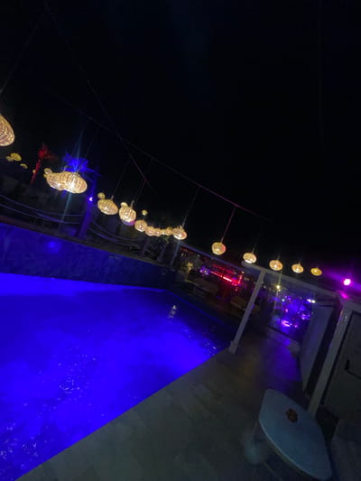 The image, likely taken at night in Mechouar Kasbah, Morocco, shows a vibrant scene with a strong emphasis on lighting. A string of decorative lights hangs diagonally across the frame, starting from the upper right and extending down to the lower left. The lights appear to be spherical with a woven or textured design. The night sky in the background is very dark. To the right of the string lights is a rectangular pool of water illuminated with a bright, saturated blue light. In the upper right corner, a terrace-like structure with various lighting, including some purple light, provides a backdrop to the scene. The area beyond the pool and below the string lights is darker, but there are hints of buildings and foliage, suggesting an urban environment.