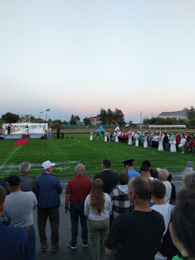 An outdoor event is taking place on a grassy field, likely a sports stadium, with spectators watching a ceremony. The scene appears to be in Таман ауылдық округі, Kazakhstan, as suggested by the presence of national flags and banners in the Kazakh language.
In the background, a stage is set up with people in formal attire on it. A banner behind the stage reads "АК БИЛГЕ - 2026" and features various graphics, including Olympic-style figures. To the right of the stage, a group of people, some in traditional Kazakh clothing, are lined up. Several flags, including the flag of Kazakhstan, are planted in the ground. A sound system with speakers is also visible.
The foreground shows a crowd of spectators from behind, facing the event. They are varied in age and attire, with some wearing casual clothing and others in more formal wear. The lighting suggests it is late afternoon or early evening. The overall atmosphere seems to be one of community gathering and celebration.
