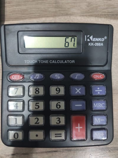 A black Kenko KK-268A calculator with a display showing "67" is on a wooden surface. The calculator has various buttons, including numerical digits, mathematical operations, and function keys like "OFF", "ON/C", "MRC", "M-", and "M+". The calculator is positioned at a slight angle, with the top-right corner closer to the viewer. The wooden surface appears to be a table or desk, with a visible grain pattern. There is no indication of the calculator's location in Yalangach, Uzbekistan.