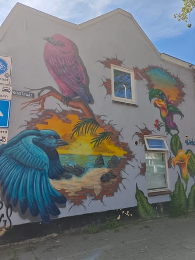 The image shows a mural on the side of a building located on Annastraat in Tilburg, Netherlands. The mural depicts two birds and a tropical landscape. On the left side of the mural, a large pink bird is perched on a branch, and below it, a blue bird with its wings spread is in the foreground. A scenic view of a beach with palm trees and a sunset can be seen through a brick wall that appears to be broken. On the right side, there are large calla lily flowers and lush green foliage. The building has two windows visible in the mural area, one of which is open. Traffic signs are present on the left side of the building. The ground in front of the building is paved with gray bricks. The sky is blue with some clouds.