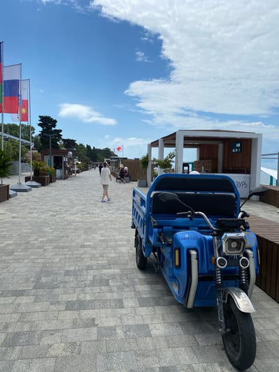 In Adler, Russia, a blue three-wheeled electric vehicle is parked on a paved promenade. The vehicle has a cargo bed and a driver's seat with two headrests. In the background, several flags are displayed on poles, including the Russian flag. A white building with an open structure and a digital display showing the time is visible further down the promenade. People are walking along the promenade, some are sitting at outdoor cafes, and others are standing near stalls. A red cross flag is also visible. The sky is blue with scattered clouds.