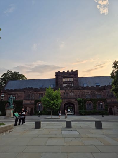 The image shows a wide-angle, eye-level view of the exterior of Firestone Library at Princeton University, located in Princeton, New Jersey, United States of America. The building, constructed of dark red stone, features Gothic architectural elements such as pointed arches, crenellations, and ivy-covered walls. A large archway in the center of the building leads to an inner courtyard. The sky above is a soft gradient of blue and yellow, suggesting either sunrise or sunset, with a few scattered clouds.

In the foreground, a paved courtyard is visible, with several dark, rectangular stone planters. To the left, there is a statue of a historical figure on a pedestal. Two people are standing near an information kiosk in front of the statue. One person, wearing a black shirt and pants, is pointing at the kiosk, while the other, in a white and black top and black pants, looks at it. Further into the courtyard, two women are walking away from the camera towards the building.

The overall scene depicts a peaceful, academic setting during twilight.