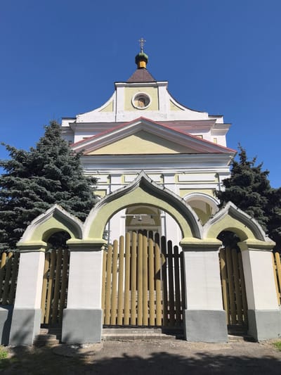 This media file is a low-angle, eye-level, full shot of a building with an ornate facade and a fence in front of it, under a clear, bright blue sky. The building appears to be a church or a similar religious structure, painted in white and pale yellow with a red roof. A small, green dome topped with a cross sits atop a dark brown spire, visible above the building's main structure. A circular icon, likely depicting a religious figure, is set into the facade. The fence is made of light brown vertical wooden planks, forming a gate in the center. Above the fence and gate, there is an arched structure, painted white and pale green, with decorative elements. Two tall evergreen trees, possibly spruces, flank the entrance, their dark green foliage contrasting with the building and sky. The sun is shining brightly, casting shadows on the ground and the fence, suggesting it is daytime, likely late morning or early afternoon. The overall impression is one of a sunny day in a peaceful, possibly rural setting. There is no visible text in the image. The location context suggests this is in the Shchorsauski Sel'ski Savet, Belarus.