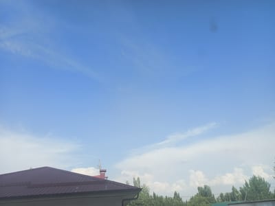 The image depicts a clear blue sky with sparse, wispy white clouds. In the foreground, the roof of a building with a burgundy metal roof is visible. A portion of a wall, likely part of the same structure, is also present at the bottom of the frame. Further in the background, a line of green trees stretches across the horizon, with a few taller, more prominent trees interspersed among them. Above the trees and on the right side of the image, a few fluffy white cumulus clouds are present. A small, dark, circular anomaly is visible in the upper right quadrant of the sky. The scene appears to be an outdoor setting in Hakkulabad, Uzbekistan, characterized by daylight and fair weather.