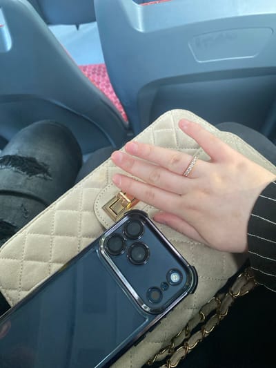 The image depicts a close-up view of a person's hand resting on a beige quilted handbag, with a dark-colored smartphone placed in front of the bag. The hand is adorned with a thin, sparkling ring. The person is wearing dark clothing, visible at the edges of the frame, including ripped jeans and a pinstripe jacket. The background suggests an indoor setting, possibly a vehicle, with blurred gray and red elements that resemble car or bus seats. The overall lighting is natural and appears to be from an overhead source. There is no indication of the location being the city of Tartu, Estonia.