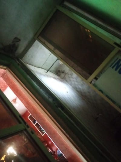 The image depicts an interior view, possibly of a shop or storage area, tilted at an angle and dimly lit. In the foreground, a green-framed window or doorway is visible, with reflections on its glass surfaces. Through this opening, a brightly lit area with pink walls and white cubbies can be seen. Some objects are present within these cubbies, appearing to be clothing or fabric items. The background is darker, with a tiled wall and a bright light source casting a strong illumination. Above, a ceiling is visible, with some discoloration or damage. In the upper right quadrant, a portion of a sign with blue lettering on a green background is partially visible, though the text is mostly obscured. The overall impression is one of a cluttered, somewhat neglected indoor space. The scene does not provide specific details that would allow for definitive identification of its location in Agbara, Nigeria, beyond being an interior environment.