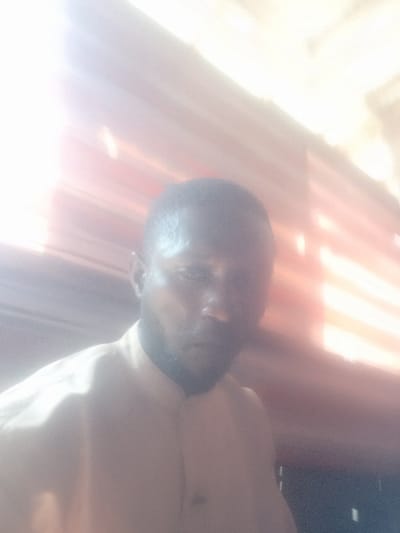 A single adult male is depicted in a close-up, blurry, and heavily overexposed image taken in Nara Unateze, Nigeria. The subject, positioned centrally, is of dark complexion with short dark hair and a beard. He is wearing a light-colored, possibly cream or light brown, collared shirt. His gaze is directed towards the camera. The background, obscured by intense glare originating from the upper left, appears to be a surface with horizontal bands of a muted reddish-orange color. No specific actions or interactions are discernible due to the image quality and framing.