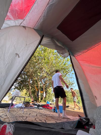 The image provides a view from inside a tent at an outdoor location within the Galil Elyon Regional Council, Israel. The tent interior features white fabric panels and a red upper section, with a patterned mat on the floor. To the left foreground, a person is lying prone on the mat near a small white-topped folding table holding a white plastic bag. Outside the tent, in bright daylight, a male stands in the mid-ground, seen from behind, wearing a white t-shirt, black shorts, and white socks; a tattoo is visible on his left calf. Further in the background, another male, shirtless and wearing light-colored shorts, appears to be walking. Various items, including clothing, are scattered on the ground and draped over tree branches. A blue cooler and water bottles are visible near the feet of the male closer to the tent. The surrounding environment consists of lush green trees under a clear blue sky.