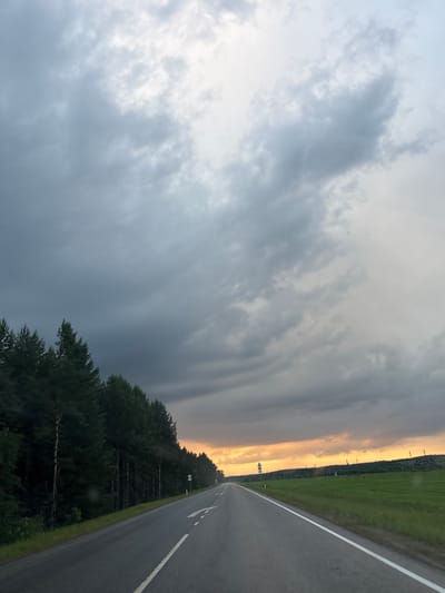 An eye-level, wide-angle perspective shot from a moving vehicle on a paved road in Malyi Kaskasal, Russia. The road, with a white line marking the lanes, curves to the left in the foreground and extends into the distance. On the left side of the road, a dense line of dark green coniferous trees creates a natural border, with their tops reaching towards the sky. On the right side of the road, a bright green, open field stretches out, bordered by distant rolling hills and a line of sparsely placed utility poles. The sky is dominated by heavy, grey, and dark clouds, with a bright band of orange and yellow light peeking through on the horizon, suggesting either sunrise or sunset. The overall atmosphere is dramatic, with the contrast between the dark, stormy sky and the illuminated horizon. There are no people visible in the scene.
