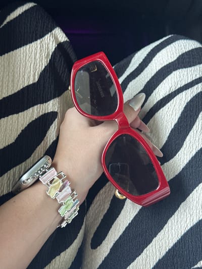 A close-up, overhead shot shows a person's right hand holding a pair of red-framed sunglasses. The hand has long, light-colored fingernails and is positioned over a black and white striped fabric, possibly a garment or upholstery. A silver bracelet with colorful pastel rectangular accents is worn on the wrist. The sunglasses have dark lenses and a brand name "JACQUEMUS" is visible on the inside of the frame. The overall scene is neutral, with no discernible context for location or specific activity. No information about Qingcheng District, China is present in the image.