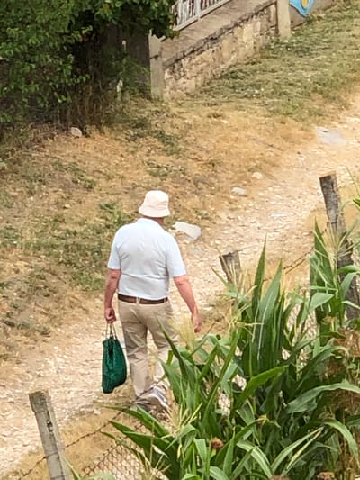 An elderly man wearing a light-colored polo shirt, khaki pants, and a bucket hat walks away from the camera along a dirt path. He carries a green mesh bag in his right hand. The path is bordered by dry grass, sparse green vegetation, and a fence made of wire and wooden posts. In the foreground, a cornfield with tall green stalks is visible. In the background, a stone wall with a railing runs along a slope covered in dry grass and some greenery. The setting appears to be a rural area, potentially within or on the outskirts of Calfa, Moldova.