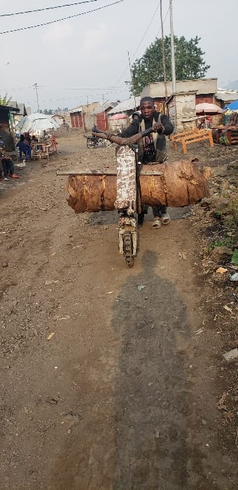 An outdoor scene in Buzove, Democratic Republic of the Congo, depicts a male individual pushing a heavy-duty, two-wheeled transport device along an unpaved dirt path. The individual, dressed in a dark long-sleeved shirt and light-colored sandals, grips the handlebars of the device with both hands, looking directly forward. The device, appearing to be a custom-built pushcart, features a white, rust-stained frame and a single front wheel. Strapped horizontally across the front of the device are two large, cylindrical bundles, approximately one meter in length each, composed of tightly packed, dried reddish-brown fibrous material.

The dirt path, light brown in color, shows evidence of foot traffic and tire marks. A long shadow of the individual and the device extends forward on the path. In the background, a dense, informal settlement is visible, characterized by numerous rudimentary structures, primarily small, single-story buildings constructed from mud, brick, and corrugated metal sheets, some with makeshift shelters or market stalls. Several other individuals are present in the background: a group seated on the left under a white tarp and another person near a parked motorcycle further down the path. Overhead power lines traverse the upper portion of the frame. Vegetation is sparse, with a prominent green-leafed tree visible on the right side of the background. The sky is overcast.