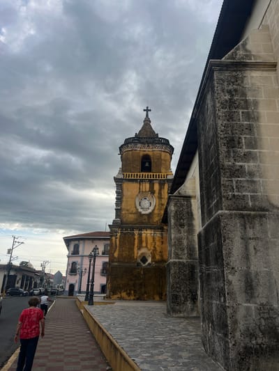 A side-angle, low-light photograph captures a portion of the León Cathedral in León, Nicaragua, under a cloudy sky. The foreground features the weathered, stone facade of the cathedral, marked by dark staining and moss. To the left, a bell tower with a yellow facade, a clock, and a bell is visible, topped by a cross. In the middle ground, a pink colonial-style building with arched windows and balconies stands beside a street lined with parked cars and utility poles. Two individuals are walking away from the camera on a patterned brick sidewalk. The street is paved with cobblestones. The overall atmosphere is muted due to the overcast conditions.