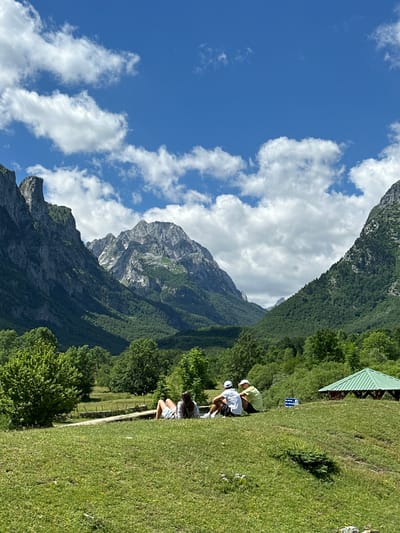 The image depicts a scenic mountain landscape in Vusanje, Montenegro. In the foreground, three individuals are seated on a grassy hill, facing away from the camera. The person on the far left is lying down, while the other two are sitting. Behind them, a green gazebo is visible, along with a sign. The middle ground is characterized by lush green trees and vegetation, leading up to a majestic mountain range that dominates the background. The sky above is a vibrant blue, dotted with white, fluffy clouds. The overall scene conveys a sense of tranquility and natural beauty.