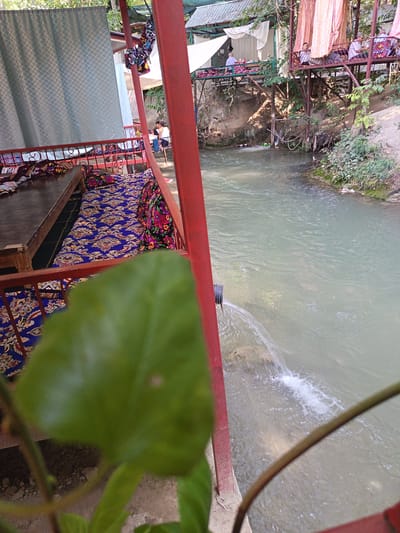 This image depicts an outdoor seating area adjacent to a flowing river, likely a cafe or restaurant setting in Langar, Uzbekistan. A wooden table is covered with a blue and gold patterned textile, suggesting seating cushions. A red metal railing separates this seating area from the river. The river flows with light brown, slightly murky water, and a black pipe is visible discharging water into it, creating a small cascade. In the background, there are elevated wooden platforms with additional seating areas, some covered with light-colored drapes. Several individuals are seated at these platforms, appearing to be patrons. The environment is natural, with trees and vegetation visible on the riverbanks and surrounding areas. The overall impression is a relaxed, possibly tourist-oriented establishment situated in a natural landscape.