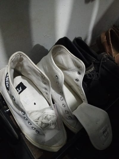 The image depicts a collection of shoes on a dark surface, presumably a shelf or rack, within an indoor environment in Al Haouafate, Morocco. In the foreground, two pairs of light-colored (likely white or off-white) canvas high-top sneakers are visible. The left sneaker contains its white laces coiled inside a clear plastic baggie, placed within the shoe's interior. The inner sole of this sneaker displays a black, stylized crown-like graphic. The right sneaker has an insole partially displaced, resting against its side. Both white sneakers feature numerous metallic eyelets for laces, which are currently unthreaded. Behind these, a pair of dark-colored (possibly black or dark blue) laced shoes, which appear to be boat shoes, are visible, with the word "WATCHOUT" partially legible on the tongue of one shoe. Further in the background, another pair of shoes, possibly brown leather, is dimly discernible. The scene is illuminated from the upper left, casting distinct shadows on a light-colored vertical surface behind the shoes. No individuals are present.