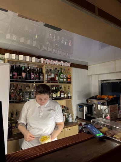 A person wearing a white polo shirt is standing behind a bar, preparing food. The bar is stocked with various bottles of liquor and other beverages. A coffee machine and a payment terminal are also visible on the counter. The scene appears to be in a bar or cafe. The image is taken from a slightly elevated angle, looking down at the person and the bar. The lighting is dim, with some light coming from above and from the coffee machine. The background is blurred, but it appears to be a shelf with more bottles.