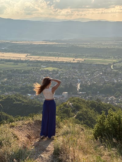 The image depicts a female figure from behind, standing on a dirt path at an elevated vantage point overlooking the city of Stob, Bulgaria, at sunset. The subject has long brown hair, illuminated by backlighting, with hands raised and holding her hair. She wears a light-colored, possibly patterned, crop top and dark blue, wide-leg pants, with sneakers visible on her feet.
In the immediate foreground and midground, dry grass, sparse green foliage, and small yellow-flowered plants cover the uneven terrain. A visible dirt path curves slightly to the right, leading down the slope. Below the subject, a dense green forested hillside transitions into the extensive settlement of Stob, characterized by numerous houses with varied roof colors, interspersed with trees and winding roads. A prominent, larger building with a flat roof is visible towards the left side of the town.
Beyond the town, the landscape flattens into a patchwork of agricultural fields, some appearing golden, others green. A road snakes through these fields. In the far distance, multiple layers of hazy, blue-grey mountains stretch across the horizon. The sky above is partially cloudy, with warm, golden light from the setting sun casting long shadows and illuminating the scene from the right, particularly highlighting the subject's hair.