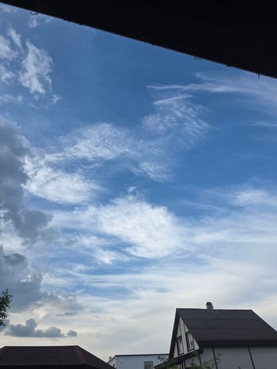 The scene depicts an outdoor environment in Brilliant, Russia, under a partially clouded sky. The upper portion of the image is dominated by a deep blue sky with numerous elongated, thin, white cirrus clouds arranged horizontally and diagonally. In the mid-left area, denser, darker grey cumulus clouds are present. The topmost edge of the frame is occupied by a dark, linear architectural element, possibly a roof overhang or gutter, from which several small, dark, thin objects (likely wires or fasteners) are suspended. In the lower-right quadrant, a residential structure is visible, featuring a steep, dark brown corrugated metal roof with a chimney at its apex. The house's exterior walls are clad in light-colored siding, accented by dark trim around gable ends and windows. Green foliage, likely tree branches, partially obstructs the view of this house and a dark-tiled roof in the lower-left. No people are visible. The scene is static, devoid of apparent actions or interactions.