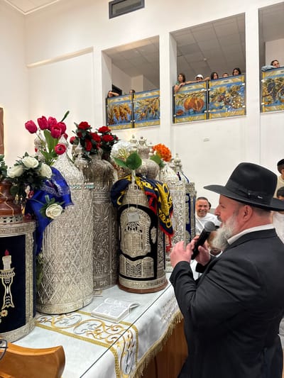 The image captures an indoor religious ceremony in Beer Yaaqov, Israel. In the foreground, a bearded man wearing a black hat and black suit holds a black microphone, speaking towards an unseen audience to his left. He stands beside a table draped with an embroidered white and gold cloth, which holds at least five ornate silver-cased Torah scrolls (tikim). Several scrolls are adorned with colorful sashes, including blue and a yellow-and-black patterned fabric. Various floral arrangements, featuring red tulips, red roses, white roses, and orange flowers, are placed among the scrolls. An open religious book lies on the table in front of the scrolls. In the background, partially visible behind the speaker, a smiling man in a white t-shirt looks forward. Above the main floor, a women's gallery features decorative panels depicting the Seven Species, behind which several women and children are visible, observing the scene from above. The room features white walls, ceiling tiles, and a rectangular ventilation opening.