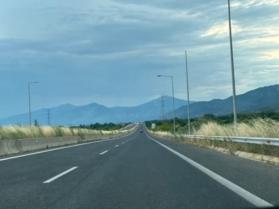 The image displays a multi-lane asphalt highway in Koptero, Greece, viewed from the perspective of a vehicle moving forward. The road features white dashed lane markers and solid white lines on the shoulders. To the left, a concrete barrier is visible, partially obscured by tall, dry, light-colored grass. To the right, a metal guardrail with similar vegetation borders the road. Multiple tall, slender street light poles extend vertically along both sides of the highway. In the midground, a dark-colored sedan travels away from the viewer in the left lane. A prominent high-voltage lattice tower is situated on the left side, with another partially visible further down the road on the right. A small white road sign with symbols is present on the right shoulder behind the guardrail. A range of distant mountains, appearing blue due to atmospheric haze, forms the horizon under an overcast sky dominated by layered gray and light blue clouds. No people are discernible.