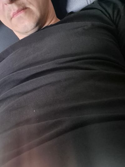 The image shows a close-up of a person's chest and neck area. The person is wearing a black t-shirt, which is wrinkled in several places, indicating it is likely made of a soft fabric. The lighting suggests the image was taken indoors, possibly in a dimly lit environment, as the black t-shirt is the dominant element and appears to absorb most of the light, with some lighter areas reflecting illumination. The bottom left corner shows a hint of skin, possibly the chin and neck, with some stubble visible. The background appears to be a textured surface, possibly upholstery from a seat. The image is framed tightly on the subject's torso and the lower part of their face, with no other elements to provide context about the surroundings or activity. There is no information in the image that can be used to determine its location in Pabradė, Lithuania.