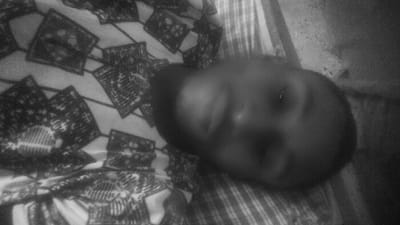 A low-light, black and white photograph, oriented approximately 90 degrees clockwise, depicts an individual lying supine in an indoor setting in Rijiyar Zaki, Nigeria. The subject's head and upper torso are visible. The individual is dressed in a garment with a distinct pattern of dark, geometrically ornate squares on a lighter background, which also covers the surface beneath their torso. A separate striped fabric is visible beneath and to the right of the subject's head. The subject's face is blurred due to low illumination, yet open eyes and basic facial features are discernible. The background beyond the immediate sleeping surface is dark and indistinct, consistent with a poorly lit interior.