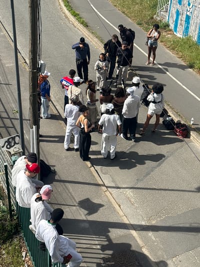 An overhead, high-angle view captures a group of approximately fifteen to twenty people gathered on a sunny street and sidewalk in Charenton-le-Pont, France. The scene suggests an outdoor production or filming activity, with several individuals holding professional cameras on tripods.
Many subjects are young adults dressed in casual clothing. Some wear all-white outfits, while others sport various streetwear styles. Notable clothing includes a blue basketball jersey with "BULLS" and the number "23" visible, a camouflage-patterned jacket, and several baseball caps in red, pink, and white, as well as a black beanie. One woman wears a white tank top and denim shorts, holding a camera.
The group is dispersed across an asphalt road and a concrete sidewalk, bordered by a grassy bank and a metal fence on one side, and a concrete wall with blue and white graffiti on the other. Long, distinct shadows indicate bright sunlight, suggesting a clear day during the late morning or early afternoon.
In the foreground, two individuals are seated on a green metal bench, both dressed in white. One holds a red smartphone. Behind them, on a concrete surface, a stylized black snake graphic is visible. Utility poles with wires run across the upper part of the frame. Scattered on the ground near some individuals are bags, including a red backpack and a black duffel bag. A patterned manhole cover is visible on the sidewalk.