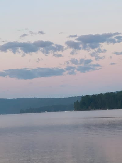 The image depicts a serene lake scene, likely at dusk or dawn, in the vicinity of Duhamel, Canada. The water is calm with a slight mist rising from its surface, creating a soft, ethereal effect. In the distance, a tree-covered shoreline forms a dark silhouette against a sky transitioning from pale blue to soft pinks. Scattered, wispy clouds are visible in the upper portion of the frame. The overall atmosphere is tranquil and peaceful, characteristic of a natural lakeside environment. No people or specific structures are clearly discernible, emphasizing the natural landscape.