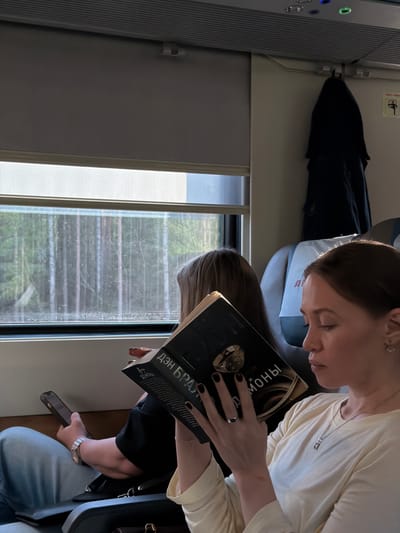 Inside a train carriage, two women are seated by a window. The first woman, with her hair pulled back, is holding a book with a dark cover and text in a Cyrillic alphabet, with the name "Дэн Браун" visible. She is wearing a light-colored top, a necklace, and rings on her fingers, and her nails are painted black. The second woman is also reading, and holding a smartphone in her hand. The train window shows a blurry view of trees passing by. On the left, there's a section of the train interior with control buttons. The overhead space contains a hanging item, possibly a jacket or bag. The location is Ушаки, Russia, based on the visible text on the book.