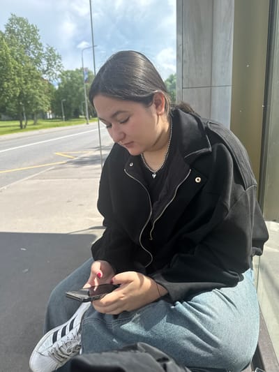 A young woman with dark hair is sitting outside, looking down at her phone. She is wearing a black jacket and blue jeans. Her fingernails are painted red. She is wearing white sneakers with black stripes. In the background, there is a street with cars, trees, and a bus stop sign. The weather appears to be sunny. The location is Yakovlevskoye, Russia.
