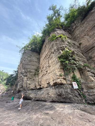 A person is standing on a rocky cliff face, looking up at the sky. The person is wearing a white top and a blue and white striped skirt, with white sneakers. They are positioned on the right side of the frame, leaning against the cliff. To the left of the person, a large, textured rock face rises up, covered in patches of green vegetation.
In the middle left of the image, there is a sign attached to the cliff. The sign is round and white with a red border and a red diagonal line through a symbol of a person falling. Text in white letters is visible below the symbol. It appears to read "CẤM XẢ RÁC".
The sky is a light blue with scattered white clouds. The lighting suggests it is daytime, possibly midday or early afternoon. The scene is outdoors, in a natural environment with a prominent rock formation. There is a green bin visible below the person on the left side of the image. The overall impression is one of natural beauty and perhaps a warning to visitors.