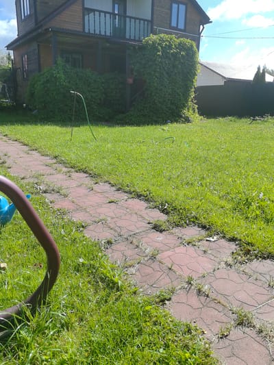 A residential property in Matchino, Russia is depicted, featuring a two-story wooden house with a covered porch and balcony. Lush green vegetation, including climbing vines on the house and a well-maintained lawn, surrounds the structure. A red brick pathway winds through the grass. A green garden hose with a spray nozzle is visible on the lawn, suggesting recent watering or preparation for gardening. The scene is bathed in bright sunlight, indicating a clear day. There are no people or animals visible in the image.