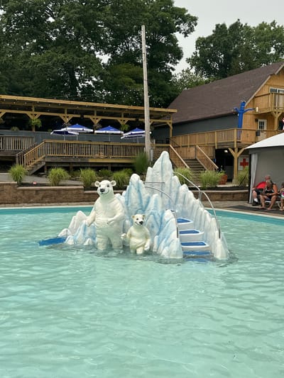 The image shows a swimming pool area at a water park or recreational facility. In the center of the pool, there is a decorative ice floe structure with two polar bear statues. A larger polar bear stands on the left, and a smaller cub is to its right. Steps are integrated into the ice floe, leading up to a platform. In the background, a multi-level wooden structure with balconies and railings is visible, possibly a restaurant or event space. Blue umbrellas are set up on a patio area in front of the wooden building. A tall, blue inflatable tube man stands on a balcony. To the right of the pool, there is a white tent with a red cross symbol on a sign, indicating a first-aid station. A few people are seated on chairs under the tent. The surrounding area is landscaped with grass and bushes, and mature trees are visible in the distance. The sky is overcast. The image appears to have been taken during the daytime in the United States, likely a warm climate given the pool setting, although the specific location in Scranton, USA, is not evident from the visual information alone.