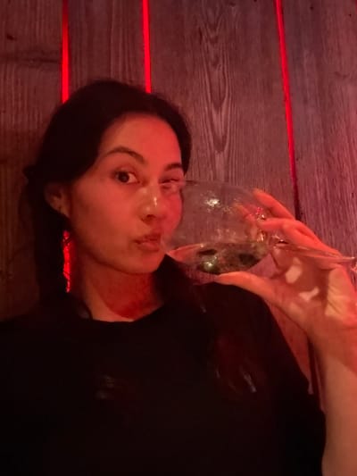 A female subject is depicted in a close-up, chest-up portrait within an interior setting in Sankt Anton am Arlberg, Austria. She possesses dark hair, styled in what appear to be two low pigtails or braids, and wears a dark-colored, long-sleeved garment. Her face is illuminated predominantly by red light, with her right eye open and her left eye partially closed. Her lips are pursed. Her right hand grasps a clear, stemmed glass, positioned at her mouth, containing a transparent liquid and discernible solid components at its base. An elongated, red-colored earring is visible on her left ear. The background consists of vertical wooden planks with visible grain. Two distinct vertical strips of intense red light are present behind the subject's head, contributing to the overall reddish illumination of the scene.