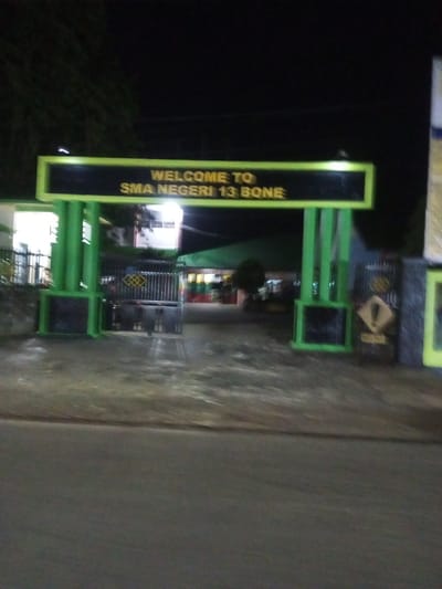 The image depicts the entrance gate of SMA Negeri 13 Bone in Walennae, Indonesia, at night. A bright yellow sign with green trim reads "WELCOME TO SMA NEGERI 13 BONE" in yellow lettering. The gate itself is made of dark metal bars and appears to be closed. Two large, bright green pillars flank the entrance, with square bases. To the left of the gate, a building with illuminated windows is visible, and to the right, a metal fence with a diamond-shaped warning sign containing a black exclamation mark within a yellow diamond is present. The ground in front of the gate is paved and wet, reflecting some of the ambient light. Trees with dark foliage are visible against the night sky. The overall scene is dimly lit, with artificial lights illuminating the entrance.