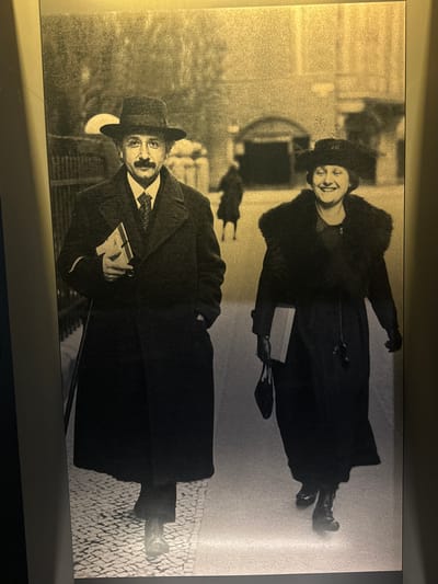 In a sepia-toned, full-length photograph, Albert Einstein and his wife, Mileva Marić, are captured walking on a cobblestone street. Einstein, on the left, wears a dark overcoat, a fedora, and a suit with a patterned tie. He carries a book in his left arm. Mileva, on the right, is smiling and wears a dark coat with a fur collar, a cloche hat, and gloves. She carries a book in her left hand and a handbag hangs from her right wrist. Behind them, blurred figures and architectural elements suggest an urban setting, possibly the city of Bern, Switzerland, where Einstein developed his theory of relativity. The lighting creates a soft, ethereal glow, emphasizing the historical and somewhat nostalgic feel of the image. The perspective is eye-level, as if the viewer is walking alongside them.