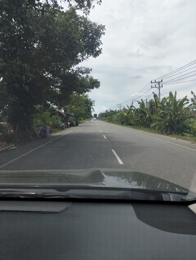 The image is a POV shot from inside a vehicle, looking out at a paved road. The road has white dashed lines dividing the lanes. On the left side of the road, there is a large tree with exposed roots and some small structures. Several motorcycles are parked along the side of the road. Further down the road, there are more trees, banana plants, and utility poles with power lines. The sky is overcast. The location is identified as Cotentung, Indonesia.
