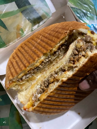 The image depicts a large, toasted sandwich, severed centrally, residing within a white cardboard takeout container. The sandwich, located in Capesterre-Belle-Eau, France, exhibits a golden-brown exterior with distinct linear grill marks. Its exposed interior layers consist of finely textured ground meat, interspersed with lighter, irregular fragments and a visible creamy white sauce. A dark-skinned thumb and index finger are partially visible on the bottom right, gently supporting the sandwich. Adjacent to the sandwich, within the container's upper left, is a crumpled transparent wrapper displaying green and yellow geometric patterns. The scene is evenly lit.
