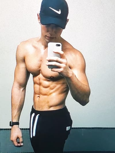 This is a medium shot, front-facing selfie of a fit man wearing a black baseball cap and black track pants. He is holding a white smartphone to take the picture. The man is shirtless, revealing his well-defined abdominal muscles and chest. He has a dark watch on his left wrist. The background is a plain, light-colored wall. The lighting is bright, suggesting it is daytime. The man's expression is neutral. The scene appears to be indoors, possibly a gym or a room with a plain wall. There is a hint of a dark tiled surface at the bottom of the image, which could be a floor or a low wall. There is no visible text in the image itself.