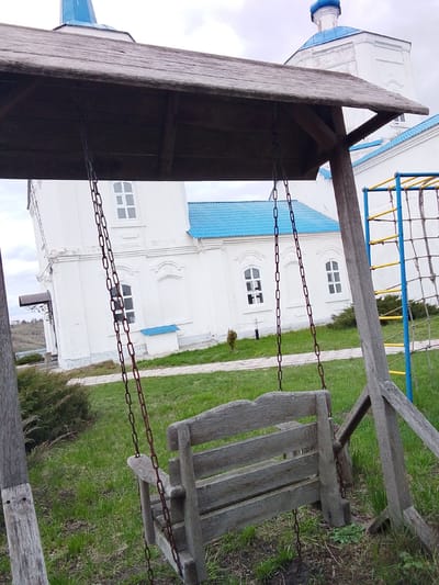 The image displays an outdoor scene in Yablonovo, Russia, featuring a rustic wooden swing set in the foreground. The swing comprises a slatted wooden bench suspended by metal chains from a wooden frame topped with a small, weathered wooden roof. The wood of the swing and its support structure appears aged.

In the midground, several white buildings with distinctive bright blue roofs and blue domes are visible. These architectural features are characteristic of Eastern Orthodox churches or monastery complexes. The buildings have multiple windows with white frames. To the right of the swing set, part of a yellow and blue metal climbing structure with rope netting indicates the presence of a playground.

The ground is covered predominantly with green grass, interspersed with some patches of bare earth and low-lying bushes. A small, sparse tree stands near one of the buildings. The sky is overcast, suggesting daytime with diffused lighting. No people are present, and no specific activity is taking place. The scene appears calm and static. There is no visible text in the image.