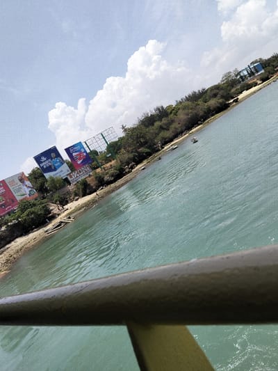 The image depicts a daytime scene looking across a body of water towards a forested shoreline in the Mkomani ward area of Mombasa, Kenya, or from Mkomani ward towards the Likoni mainland. The perspective is angled approximately 15 degrees clockwise from horizontal. In the immediate foreground, a dull olive-green metal railing, approximately 10-15cm in diameter and exhibiting minor surface wear and a single crack, extends diagonally from the bottom-left to the bottom-right. The midground consists of a teal-green body of water with rippled surfaces. Two small, light-colored boats are visible near the distant shoreline, with one appearing to hold a solitary, indistinct object or person. The background features a narrow, rocky and sandy shoreline transitioning into a dense, elevated strip of green foliage, comprising trees and bushes. Above the tree line, three large billboards are prominently displayed. The leftmost billboard is partially obscured but shows text including "OPEN" and green-on-white graphics. The central billboard advertises "BARDUI FC IN TEYU" with an image of a male football player in a blue jersey and the brand "TUPATANE." The rightmost billboard features "3X MORE," "DIESEL," and "PETROL" with red, white, and black graphics, and below it, a sign reads "LIKONI QUALITY FURNITURE." In the far right background, a building with a blue roof is partially visible. The sky is bright blue with scattered large, white cumulus clouds, indicating sunny conditions. No clearly discernible human interactions or specific actions are visible, beyond the potential presence of an individual in the small boat.