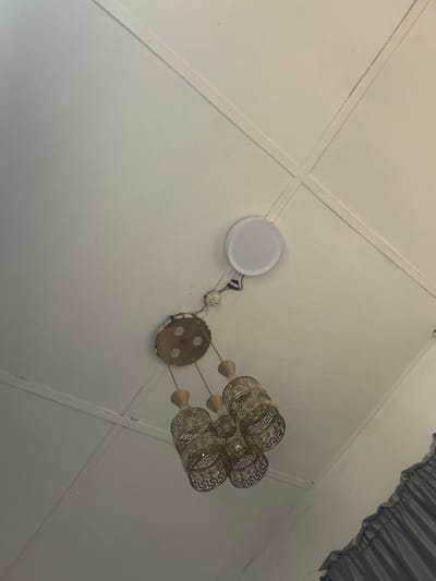 This image shows a white ceiling in an interior space located in Ilishan, Nigeria. The ceiling structure features a visible grid pattern from panels or tiles. A multi-pendant chandelier is suspended, consisting of a round, metallic base from which three cylindrical light shades hang. Each shade is gold-toned with a repeating Greek key-like geometric pattern. Adjacent to the chandelier, a white, circular, flush-mounted fixture, appearing as either a recessed light or a ceiling speaker, is installed. Exposed electrical wiring and an uncovered electrical junction are visible between the chandelier's base and the circular fixture, indicative of an exposed or unfinished electrical installation.