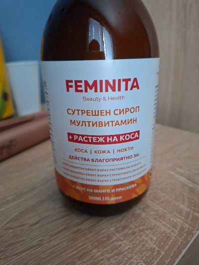 An amber-colored bottle, identified by its label as "FEMINITA Beauty & Health," stands upright on a light brown wooden surface featuring visible grain patterns. The label is predominantly white, with "FEMINITA" and key phrases in red Cyrillic text. Translated, the label indicates "Morning Syrup Multivitamin" with an emphasis on "+ Hair Growth," benefiting "Hair | Skin | Nails." The bottom portion of the label is orange with a subtle pattern and states "With Mango and Peach flavor," specifying "500ML | 25 doses." Reflections are present on the bottle's surface. In the blurred background, to the left of the bottle, a partial yellow object is visible, followed by a white, cylindrical object, possibly a cup or container. The furthest background consists of a muted blue-gray wall or surface. The specific location within the city of Selyanin, Bulgaria, cannot be determined from the provided image. No individuals are present, and no actions or interactions are depicted.
