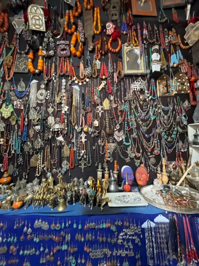 The image shows a stall in a marketplace, located at 31.329 latitude, -7.377 longitude, in Tamguinguante, Morocco. The stall displays a wide array of jewelry and trinkets, primarily necklaces, earrings, and charms, made of various materials including metal, beads, and stones. The items are arranged neatly and densely on the wall, creating a visually rich and colorful display. There are multiple small decorative figures of animals and other motifs, along with small decorative bowls and a teapot, sitting on a counter at the base of the stall. There are no people visible in the image. The stall is located in a bustling marketplace, where tourists and locals alike can purchase beautiful handcrafted items from the local artisans. The image focuses on the artistry and cultural heritage of the region, as reflected in the intricate details and vibrant colors of the jewelry.