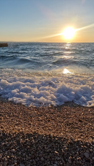 This media file captures a serene beach scene at sunset in Zadar, Croatia. The sun, a bright orb with prominent rays, hangs low in the sky, casting a warm, golden glow across the image. The sky transitions from a clear, pale blue on the left to a soft yellow as it approaches the horizon.

The scene is divided into distinct elements: the vast expanse of the sea, the foamy edge of a wave, and a pebble beach. The sea exhibits gentle ripples, reflecting the sunlight and creating a shimmering effect. In the foreground, a wave has receded, leaving behind a thick layer of white foam that clings to the shore. This foam is textured with numerous small bubbles, catching the light and appearing almost iridescent.

The beach itself is covered in a dense scattering of small, smooth pebbles. The warm light of the setting sun highlights the individual stones, creating a play of light and shadow across the textured surface. The pebbles are predominantly in shades of brown, gray, and beige. A hint of a wooden post is visible at the bottom left, partially submerged near the water's edge.

The overall mood is one of tranquility and natural beauty. The time of day is clearly sunset, indicated by the low angle of the sun and the warm, golden light. The weather appears calm and clear, suitable for a peaceful evening by the sea. No people or visible text are present in the image.