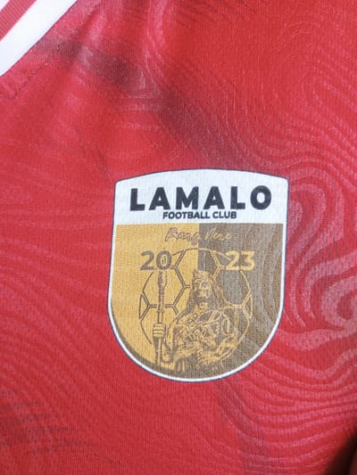The image shows a close-up of a red sports jersey with a crest. The crest is shield-shaped and features the text "LAMALO" at the top, followed by "FOOTBALL CLUB" in smaller lettering. Below this, within the shield, are the numbers "207" on the left and "23" on the right, separated by a stylized graphic that appears to be a depiction of a person holding a weapon or staff. The jersey itself has a subtle textured pattern. The crest is stitched onto the fabric. The specific location in the city of Turumame, Indonesia, is not determinable from the image.